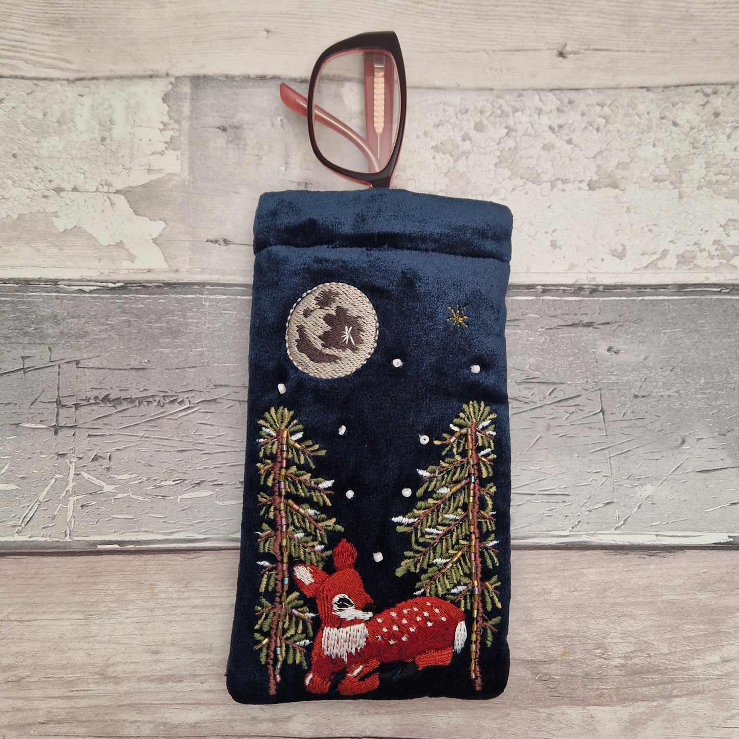 A dark navy blue velvet eyeglasses pouch with an embroidered winter forest design featuring a red deer, pine trees, a moon, and stars. A pair of eyeglasses with brown frames is resting inside the pouch, which is displayed on a rustic, light-colored wooden plank background.