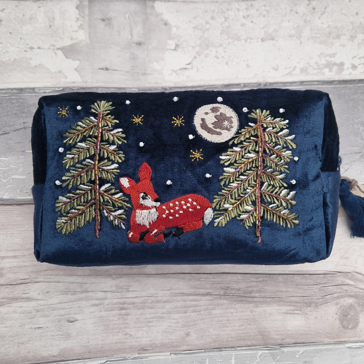 A navy blue velvet cosmetic pouch featuring an embroidered winter forest scene with a red deer, pine trees, stars, and a moon, displayed on a light wood surface.