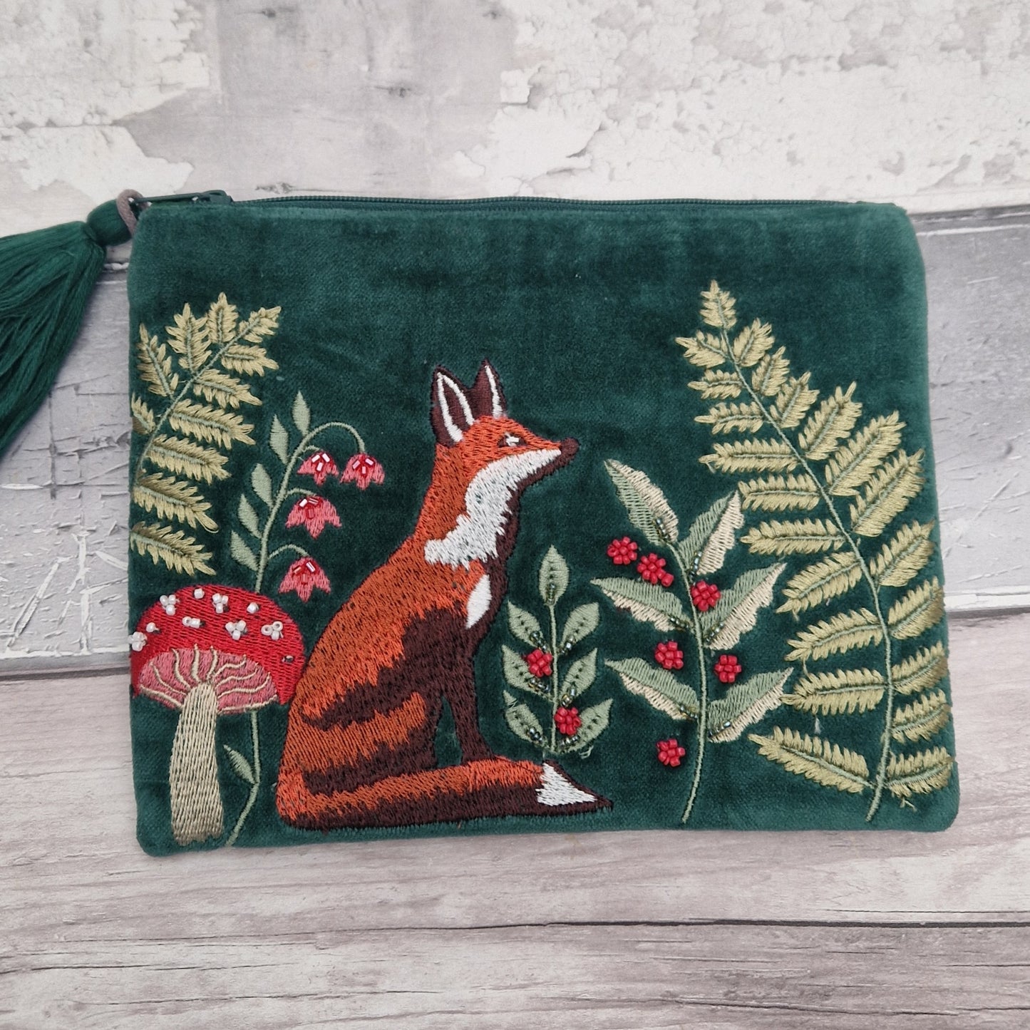 A dark green velvet zipper pouch featuring an embroidered forest scene with a fox, ferns, berries, and a mushroom, resting on a light wood surface.