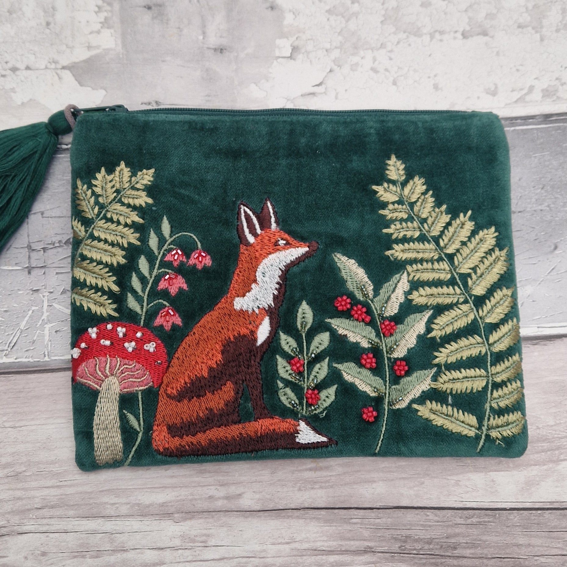 A dark green velvet zipper pouch featuring an embroidered forest scene with a fox, ferns, berries, and a mushroom, resting on a light wood surface.