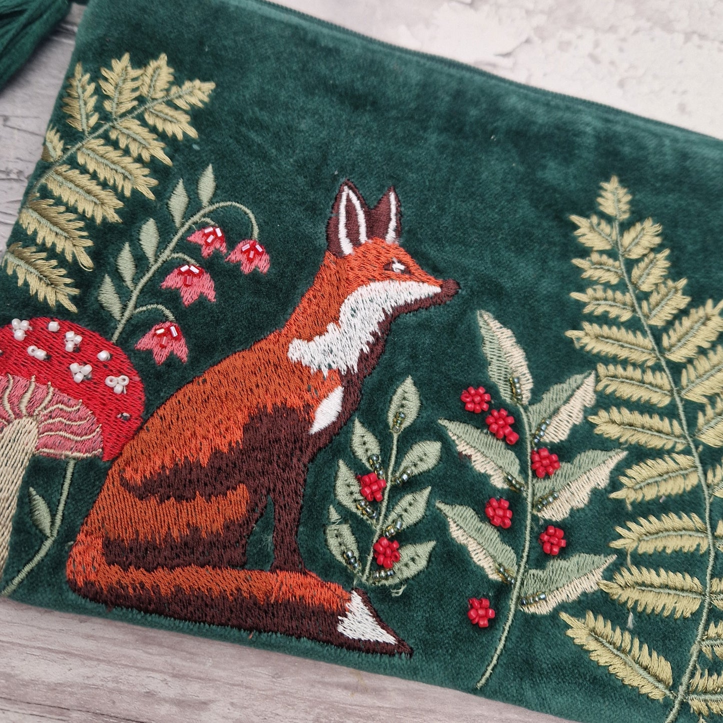 A close-up of a dark green embroidered textile featuring a fox, ferns, berries, and a red-and-white mushroom, laid on a light wood surface.