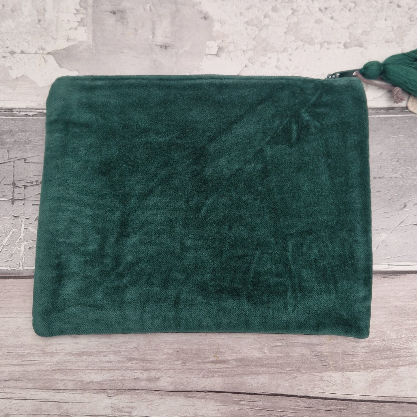 A dark green plush velvet pouch or clutch with a zipper closure and a matching green tassel, displayed on a rustic light gray wooden surface against a textured white wall.
