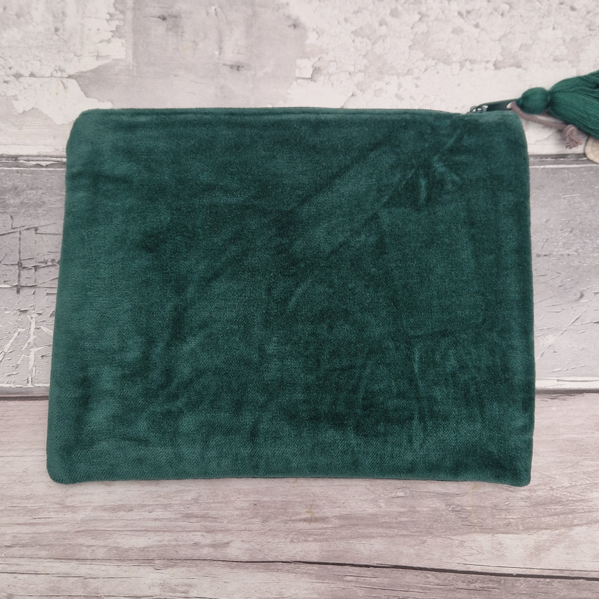 A dark green plush velvet pouch or clutch with a zipper closure and a matching green tassel, displayed on a rustic light gray wooden surface against a textured white wall.