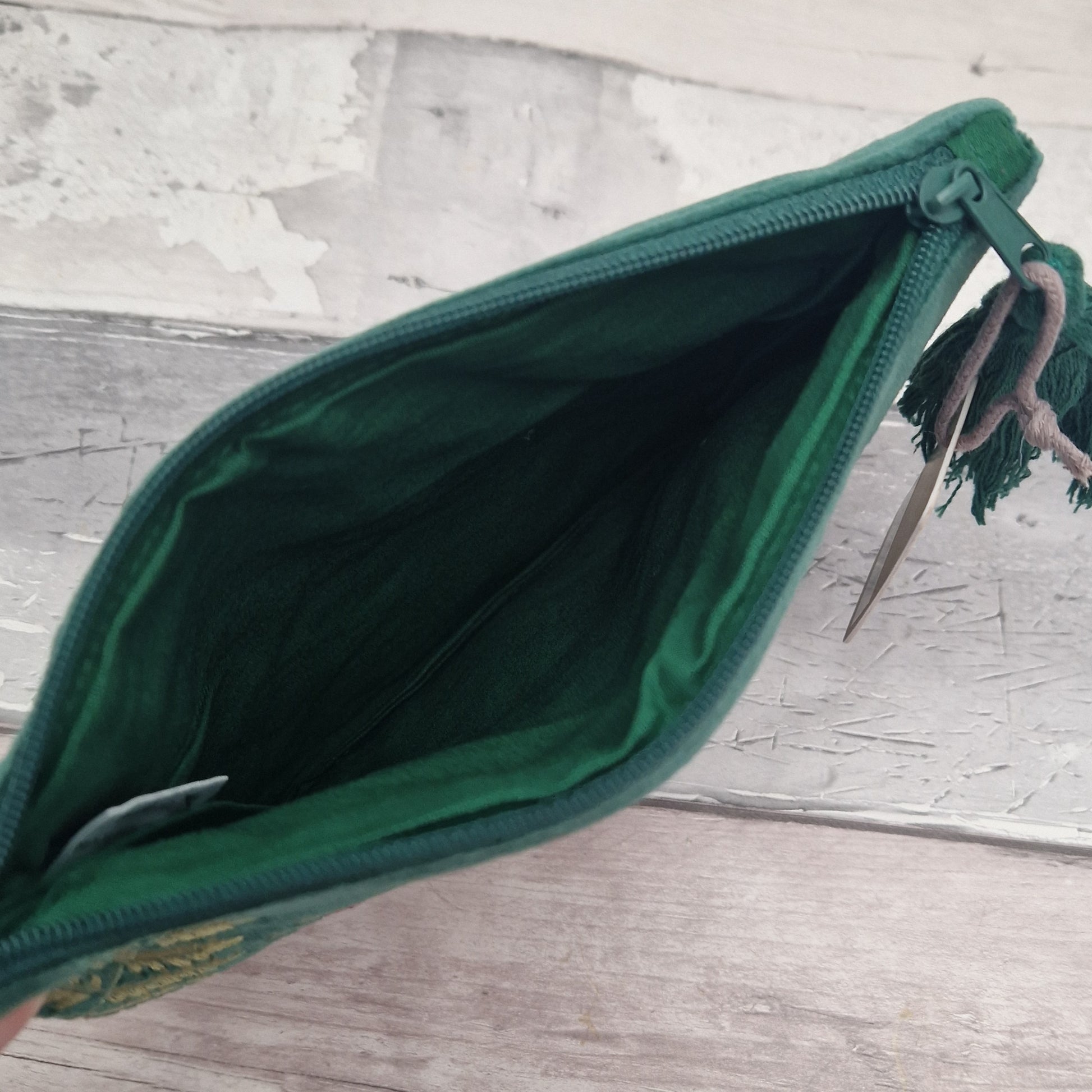A dark green velvet zippered pouch with a tassel zipper pull, shown open to reveal its interior lining, resting on a rustic light gray wooden surface.