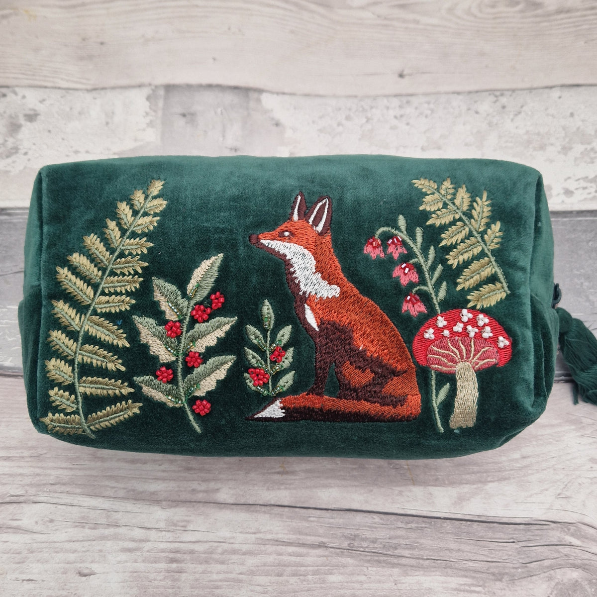 A dark green velvet pouch or cosmetic bag featuring detailed embroidery of a fox, ferns, berries, and a red-and-white mushroom, displayed on a light gray wood surface.
