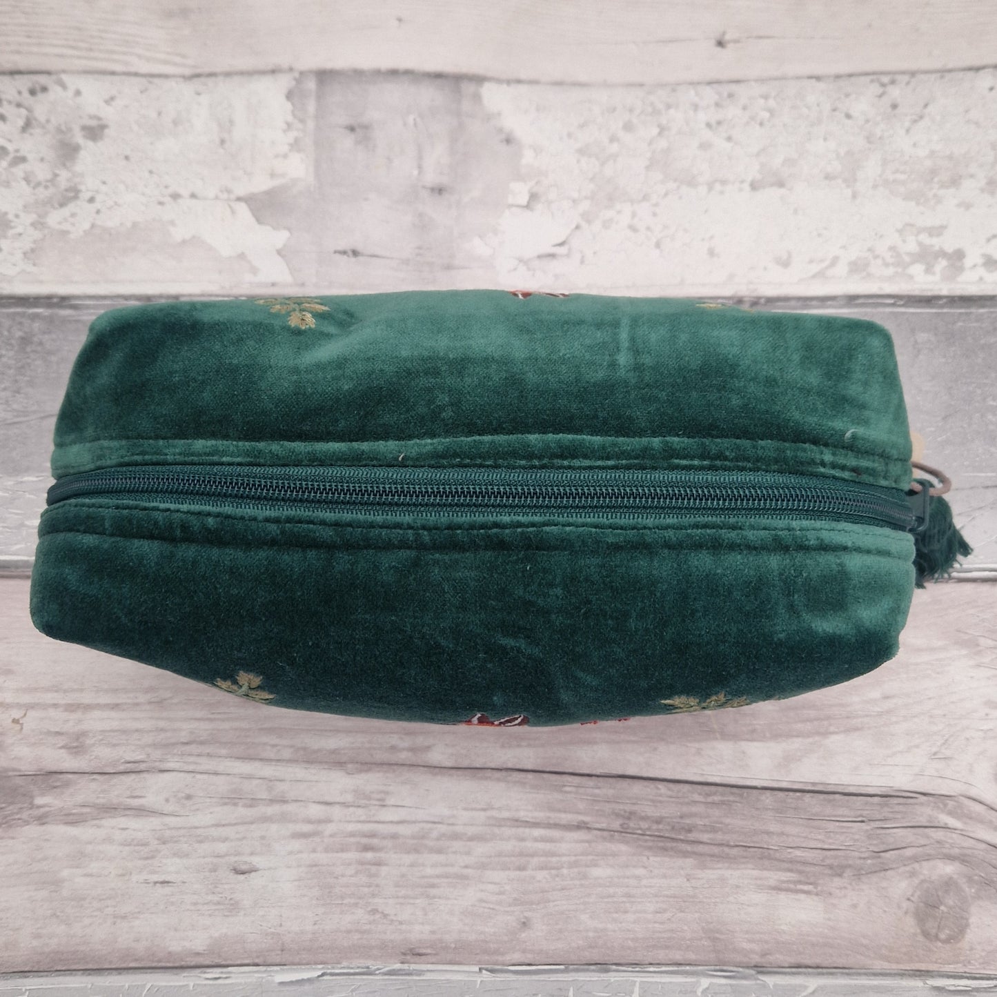 A dark green velvet zippered pouch with small embroidered floral details, resting on a rustic light gray wooden surface.