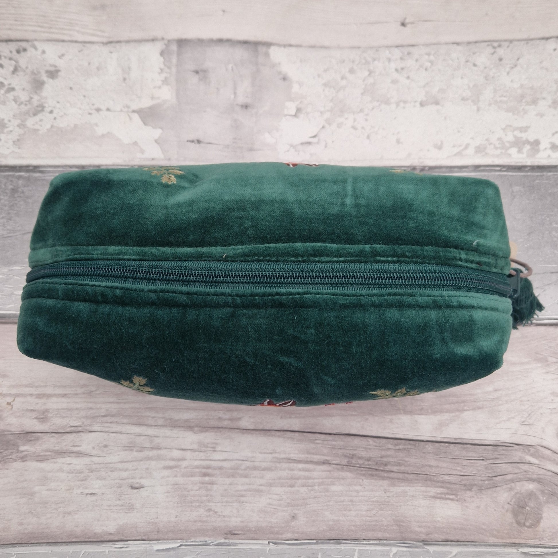 A dark green velvet zippered pouch with small embroidered floral details, resting on a rustic light gray wooden surface.
