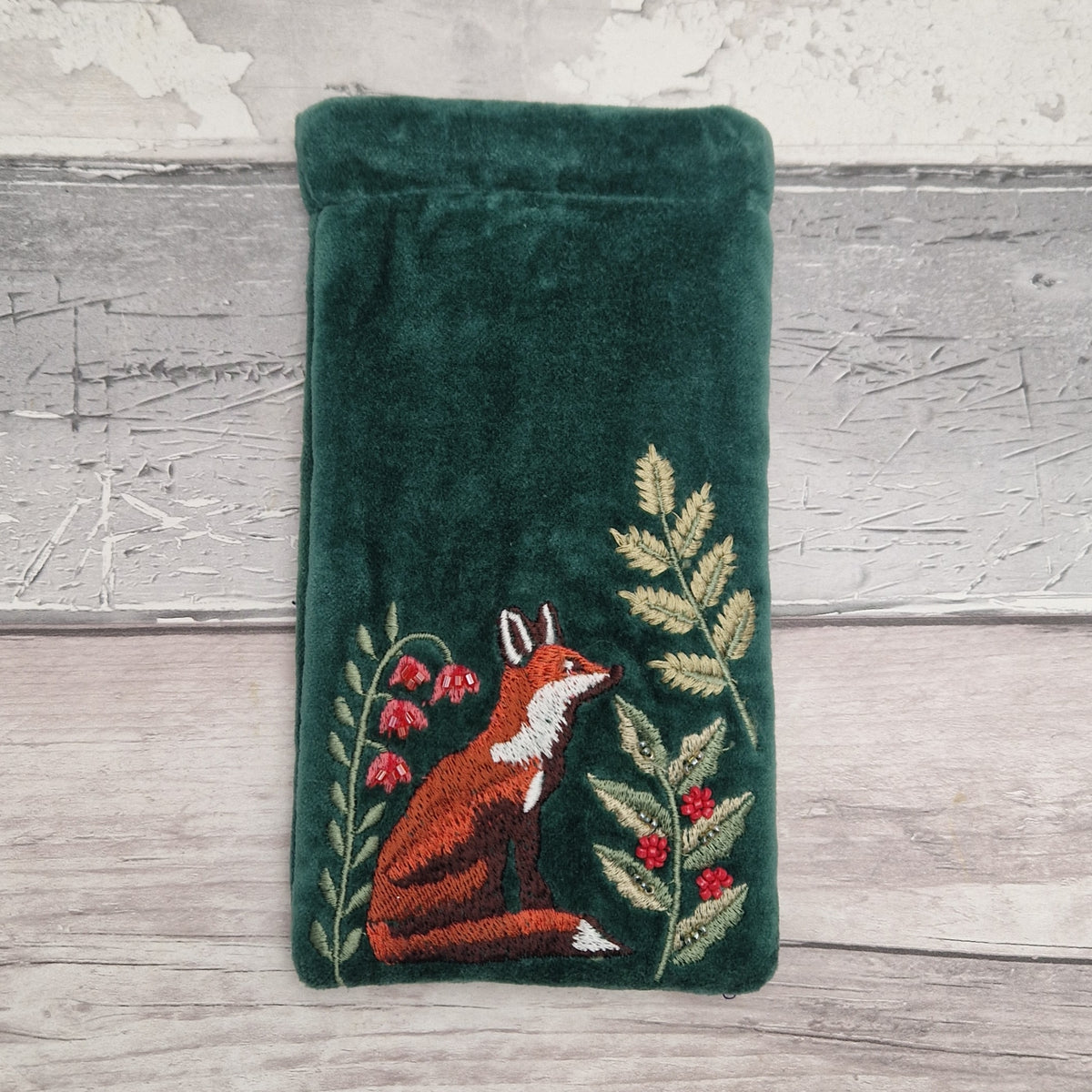 A dark green velvet pouch featuring an embroidered design of a fox, ferns, and red berries, displayed on a rustic gray wooden surface.