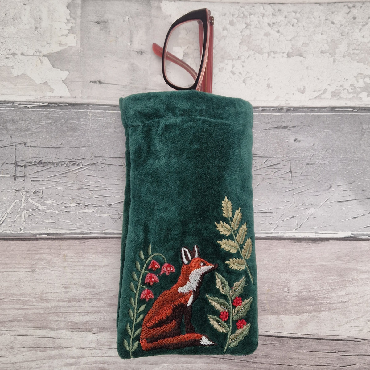 Green velvet glasses case decorated with a embroidered fox sat elegantly amongst woodland ferns. A pair of glasses are poking out of the top of the case.
