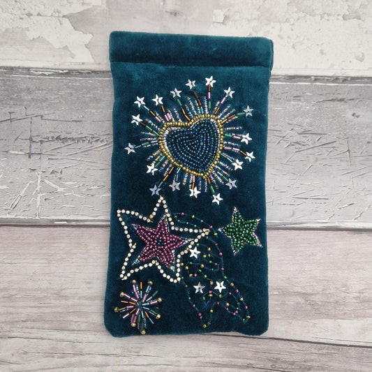 A dark teal velvet glasses case decorated with colorful beaded stars, a heart, and fireworks motifs, displayed on a rustic gray wooden surface.