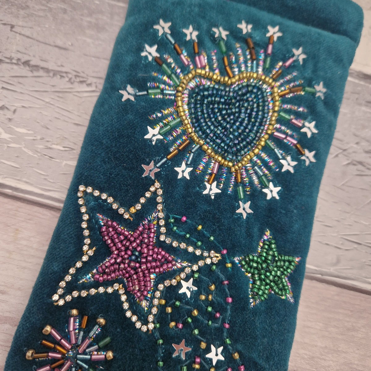 A teal velvet glasses case decorated with colorful beaded stars, a beaded heart, and festive starburst patterns, laid flat on a light wood background.