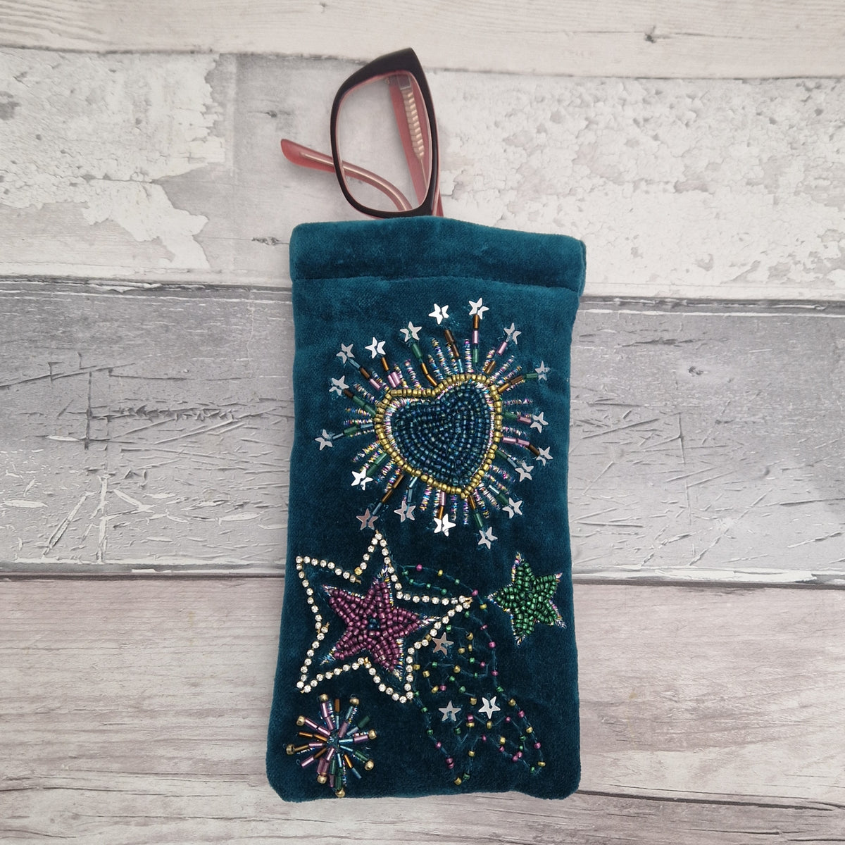 A dark teal velvet eyeglasses pouch decorated with colorful rhinestone stars and a heartburst design, shown with a pair of glasses resting inside on a rustic white-washed wood background.
