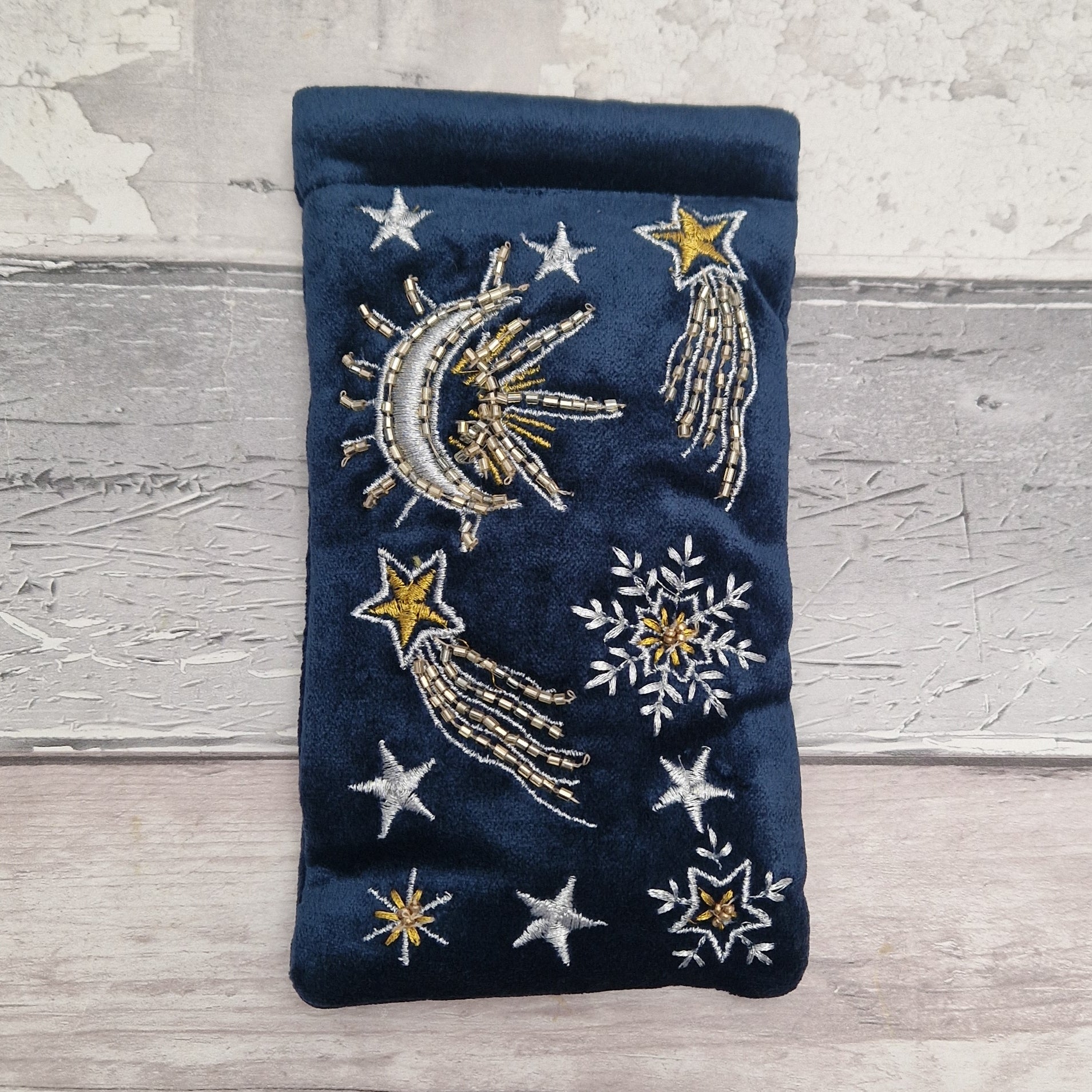 A navy blue velvet pouch decorated with embroidered celestial motifs including a crescent moon, shooting stars, and snowflakes, displayed on a light gray wooden surface.