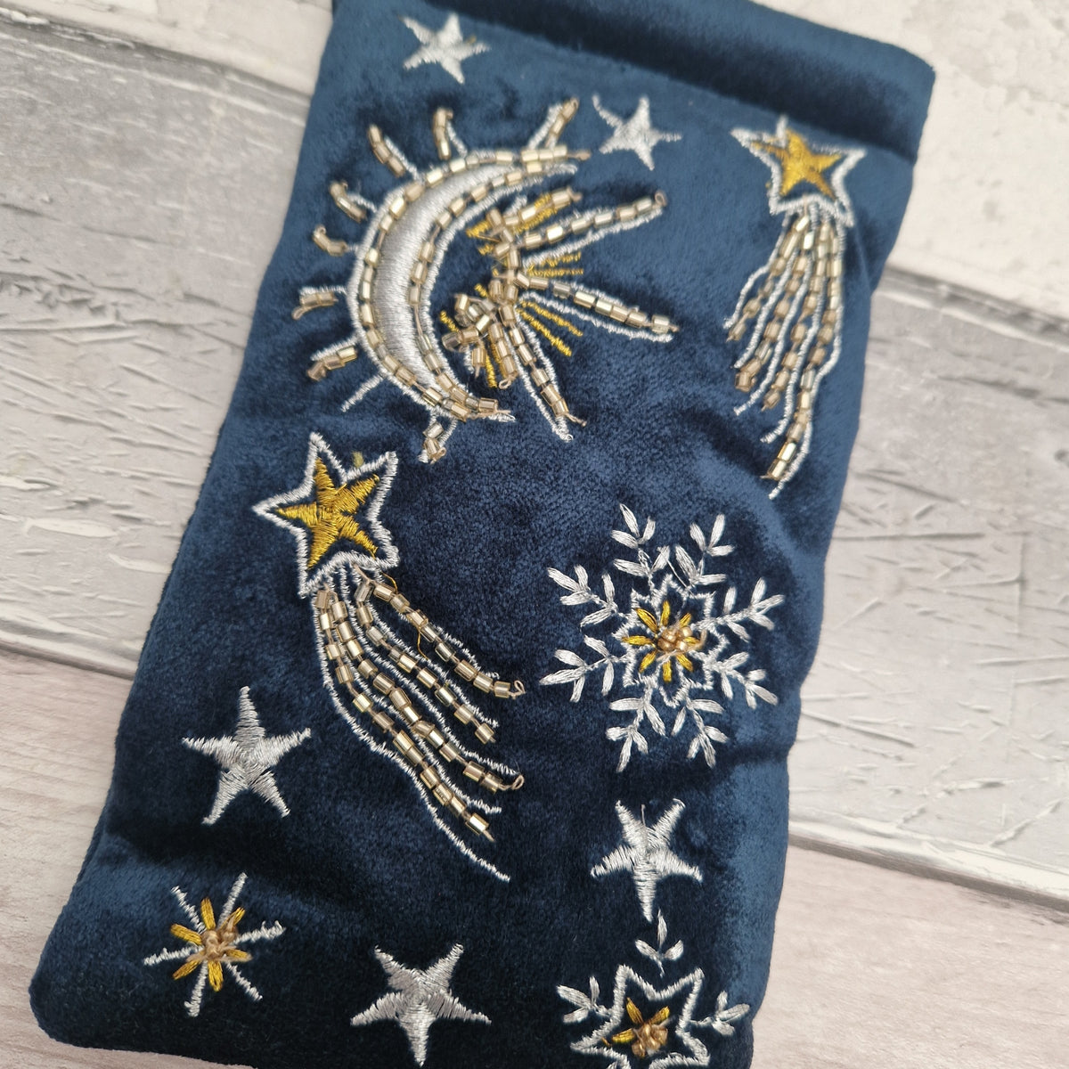 A navy blue velvet pouch decorated with embroidered celestial motifs including a crescent moon, stars, comets, and snowflakes in white and gold thread, shown on a light gray wood background.