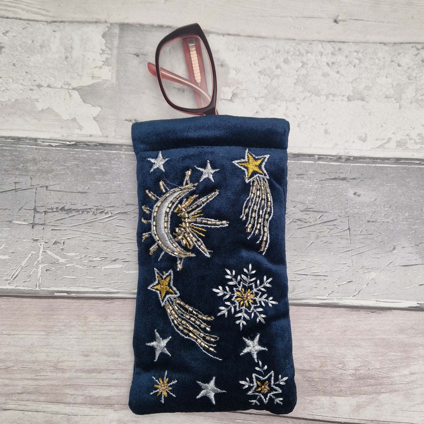 A navy blue velvet eyeglasses pouch with embroidered celestial designs including a crescent moon, shooting stars, and snowflakes, shown holding a pair of eyeglasses on a rustic white-washed wooden background.