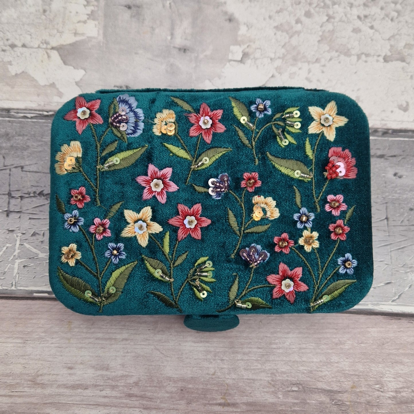 A teal velvet jewellery box featuring colorful floral embroidery in pink, yellow, blue, and red, resting on a light gray wooden surface against a textured gray wall.