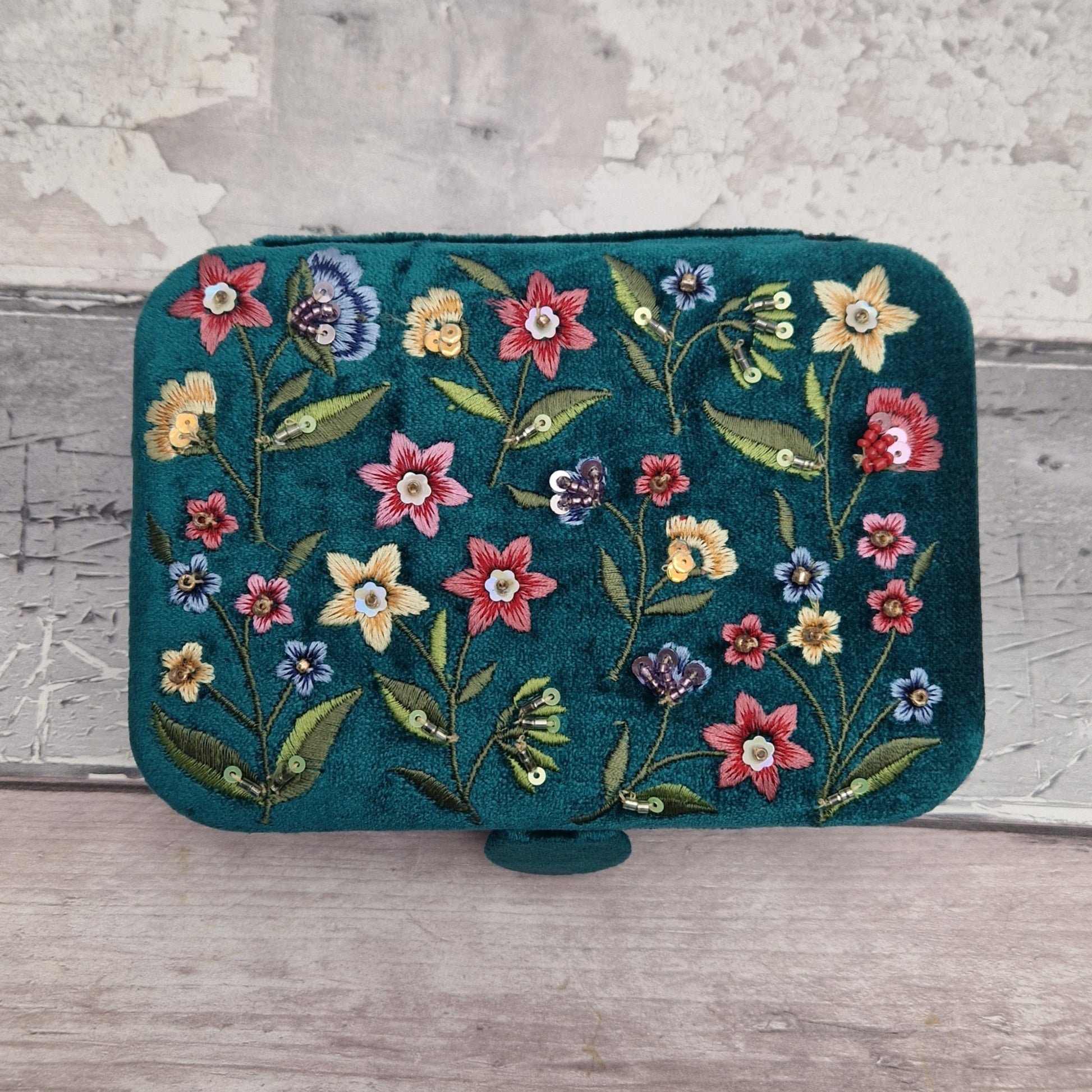 A teal velvet jewellery box featuring colorful floral embroidery in pink, yellow, blue, and red, resting on a light gray wooden surface against a textured gray wall.