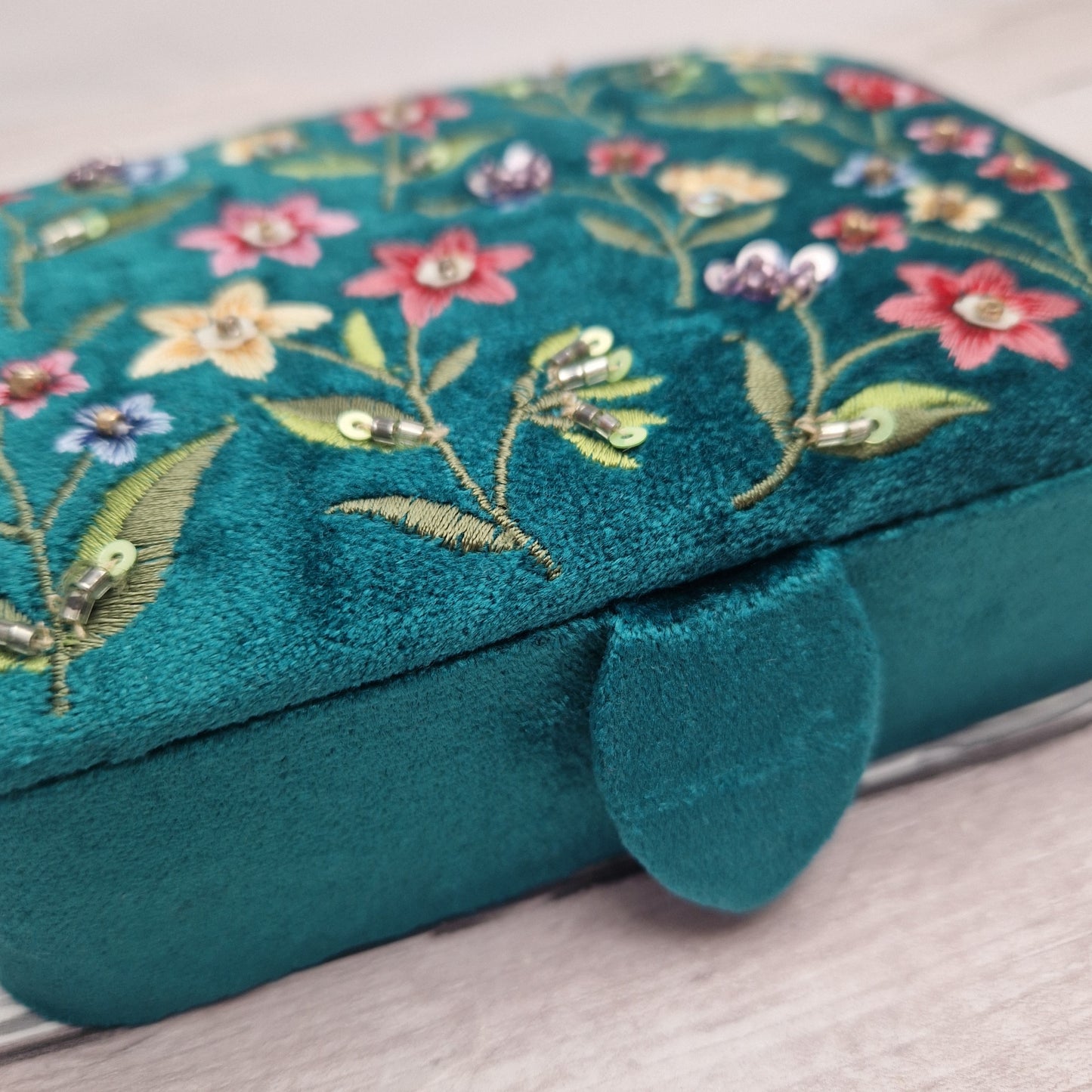 A close-up of a teal velvet jewelry box with colorful floral embroidery and sequins on the lid, resting on a light wood surface.