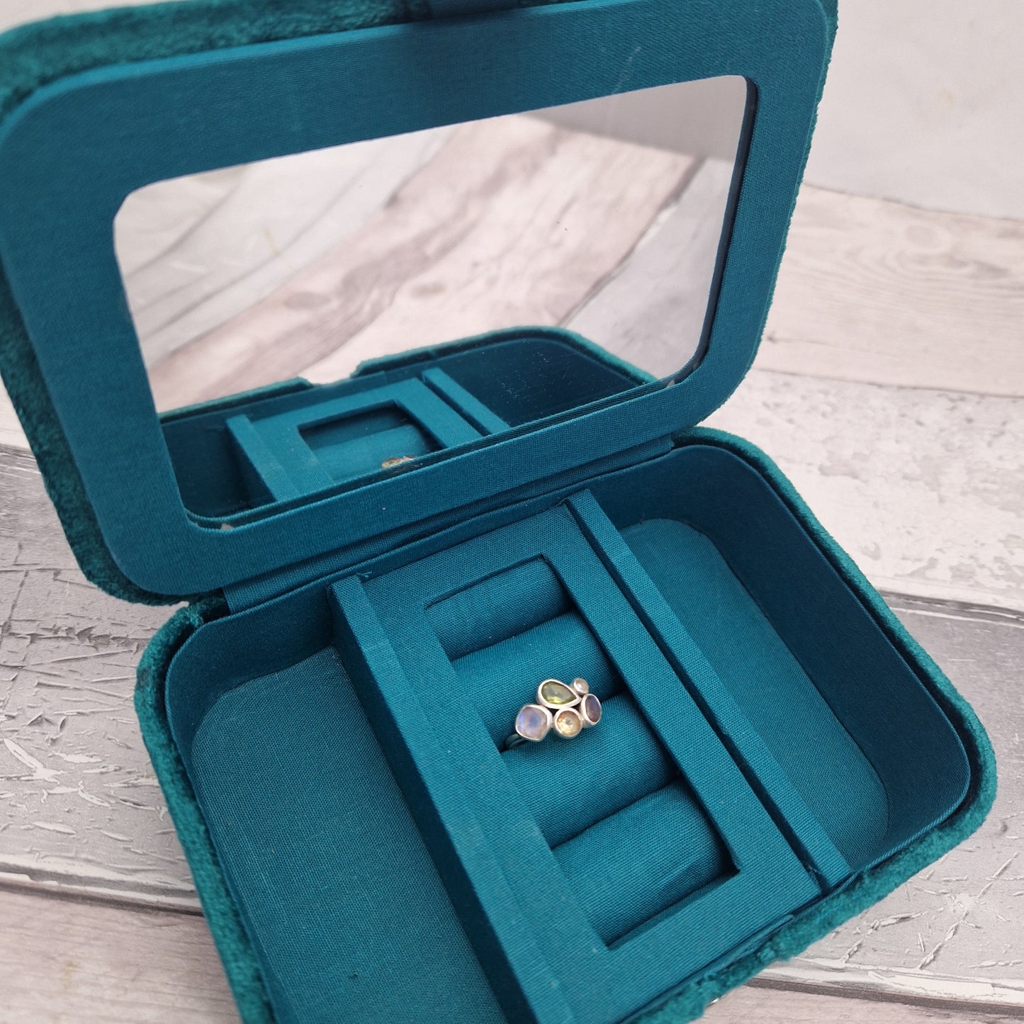 An open teal velvet jewellery box with a clear mirror in the lid, showing a small gold ring with purple and blue stones inside one of the ring slots, set on a light wood surface.