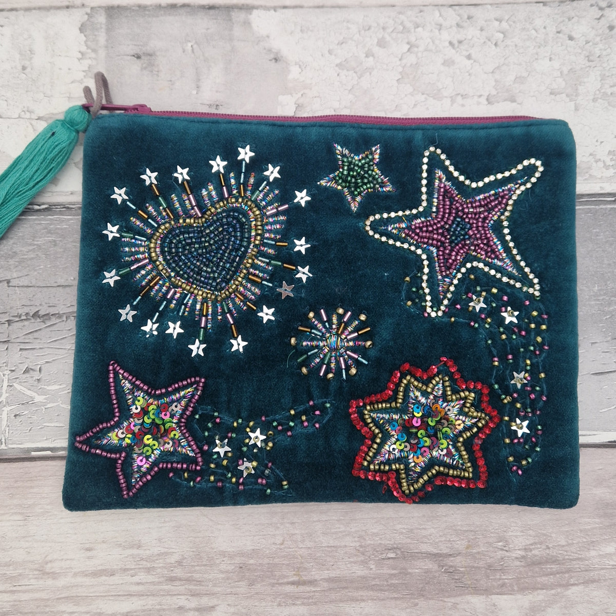 A dark teal velvet zip pouch decorated with colorful beaded stars, a beaded heart, and a teal tassel, displayed on a light gray wooden background.