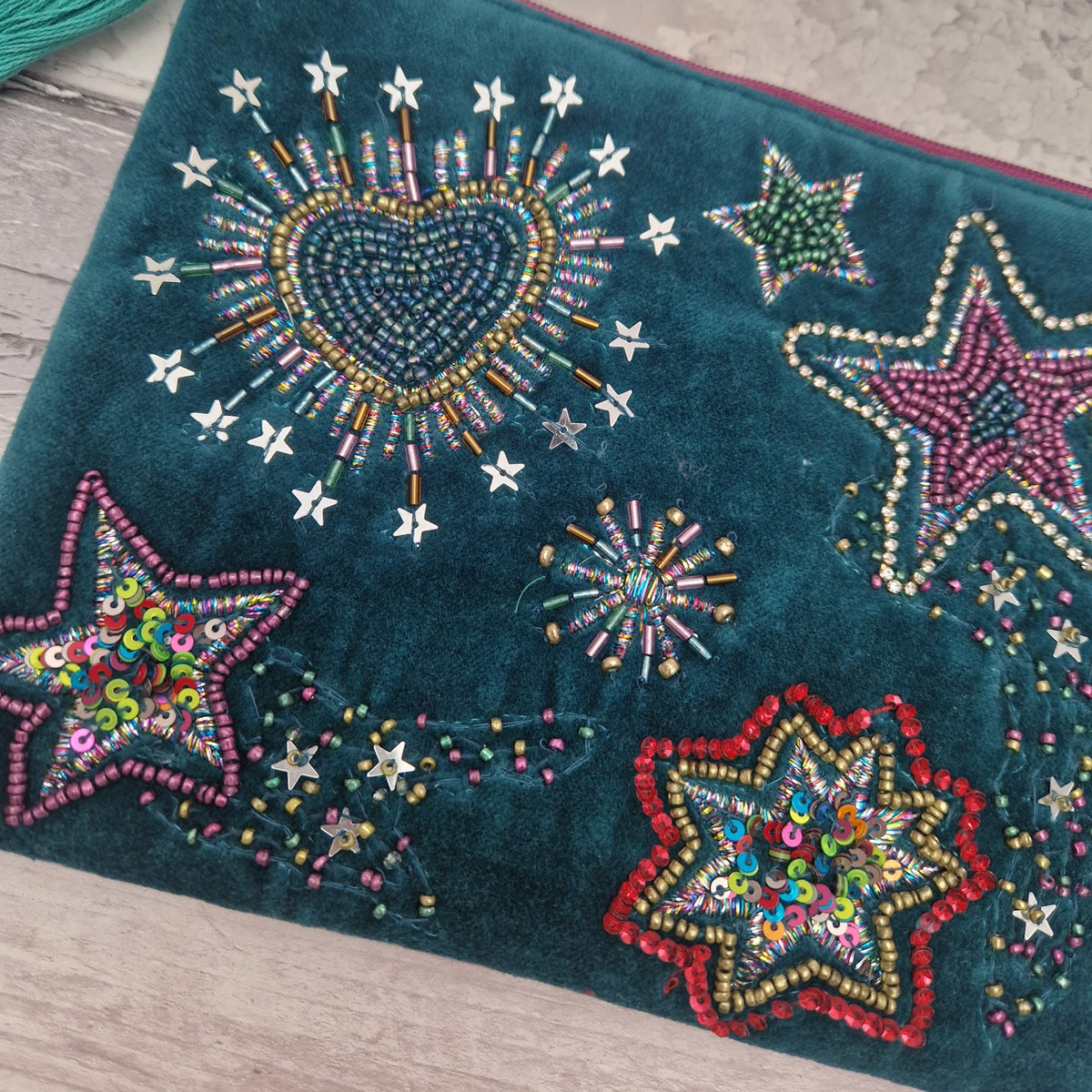 A close-up of a dark teal velvet pouch decorated with colorful beaded star and sunburst motifs on a light wood surface.