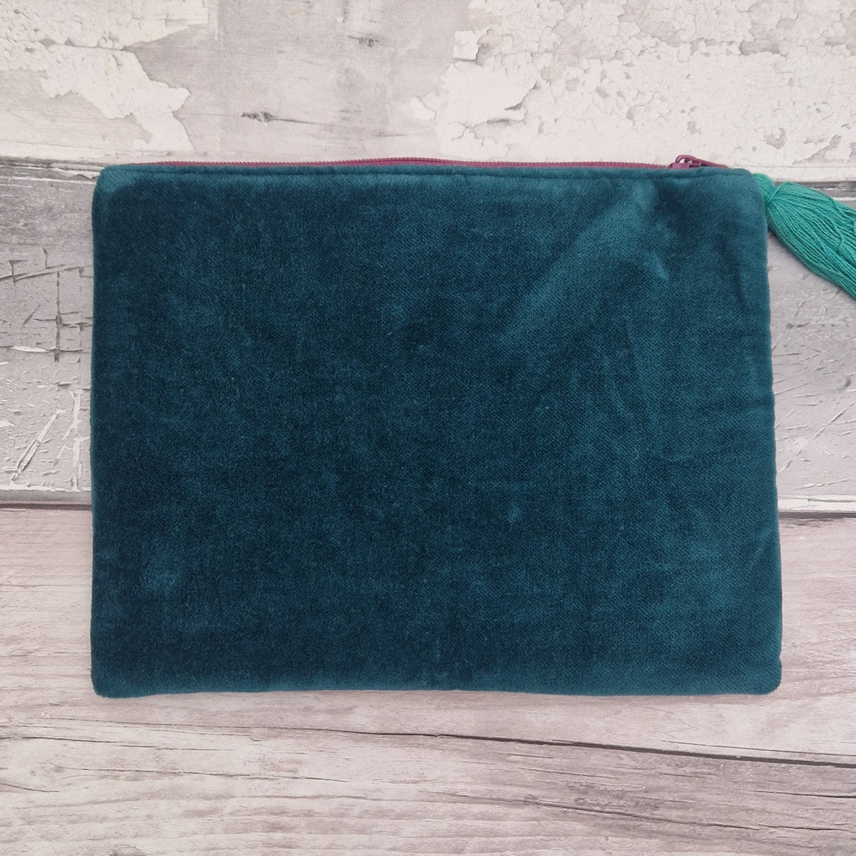 A dark teal velvet pouch with a pink zipper and a matching green tassel, displayed on a rustic light gray wooden surface.