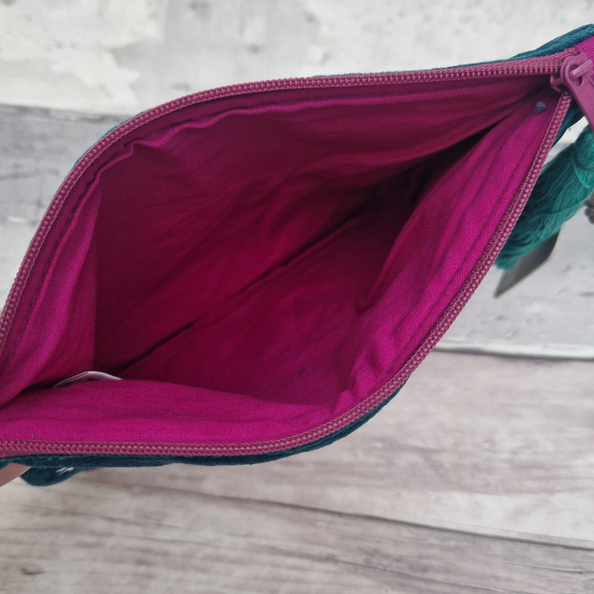 An open zippered pouch or clutch shown from the inside, with a bright magenta-pink lining and a dark teal fabric exterior visible along the edges and zipper tape, set against a light wood surface.