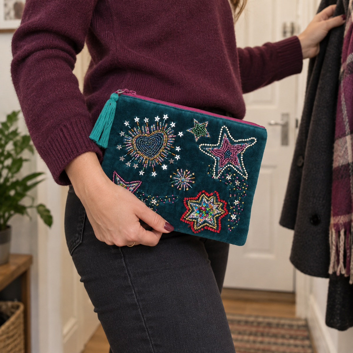 A person wearing a burgundy sweater and dark jeans holds a teal velvet zip pouch decorated with colorful beaded star and sunburst motifs. The scene is set in a home interior with a door, plant, and hanging coat visible.