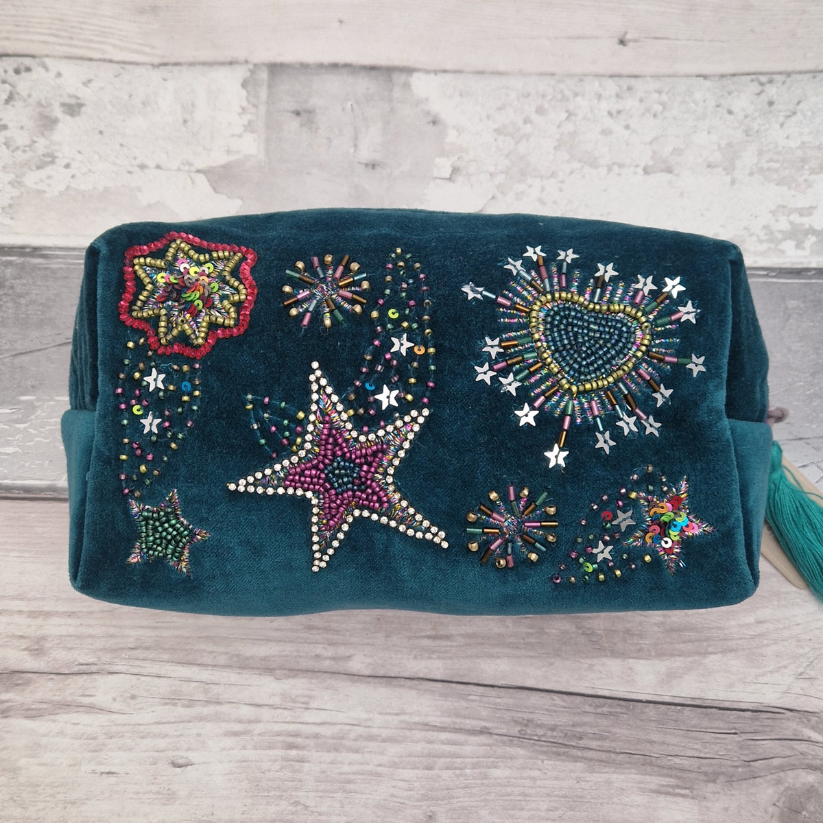 A dark teal velvet wash bag decorated with colorful beaded stars, hearts, and fireworks patterns, resting on a light gray wooden plank surface.
