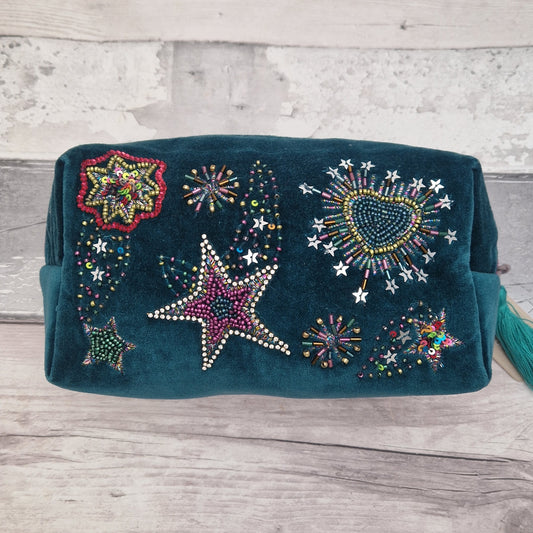 A dark teal velvet wash bag decorated with colorful beaded stars, hearts, and fireworks patterns, resting on a light gray wooden plank surface.