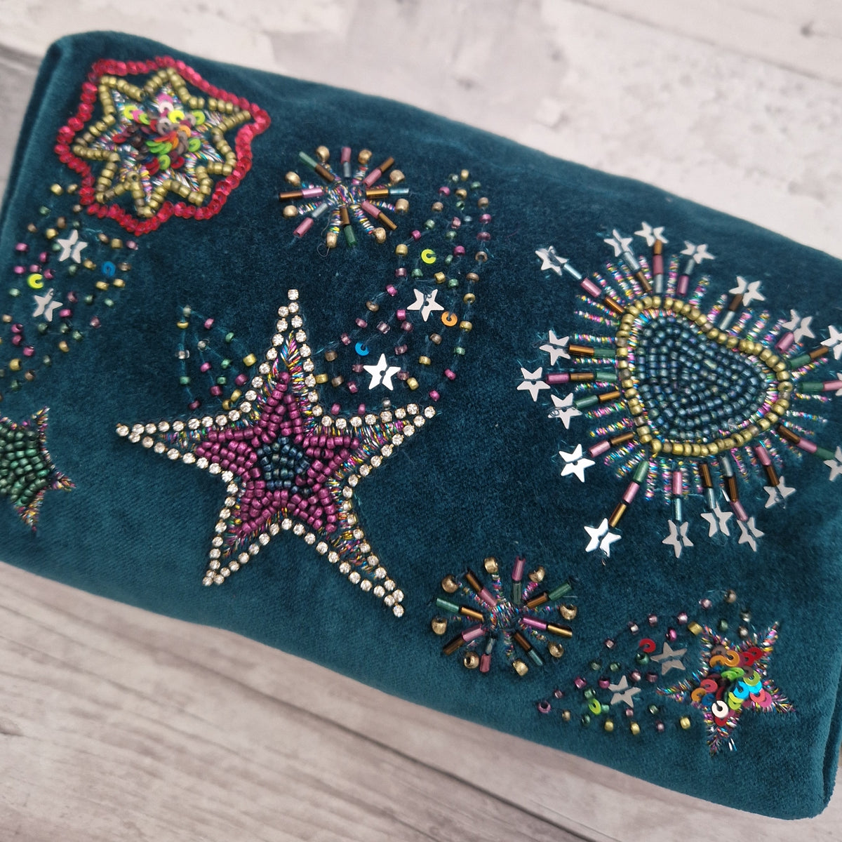 A dark teal velvet wash bag decorated with colorful beaded star, heart, and firework motifs, resting on a light wood surface.