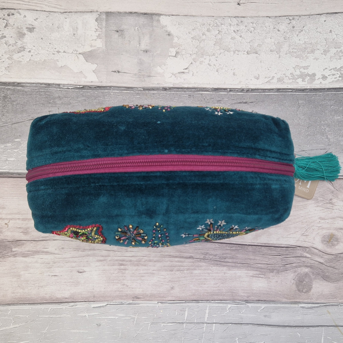 A teal velvet zippered pouch with colorful floral embroidery and a magenta zipper, resting on a rustic white-washed wooden surface.