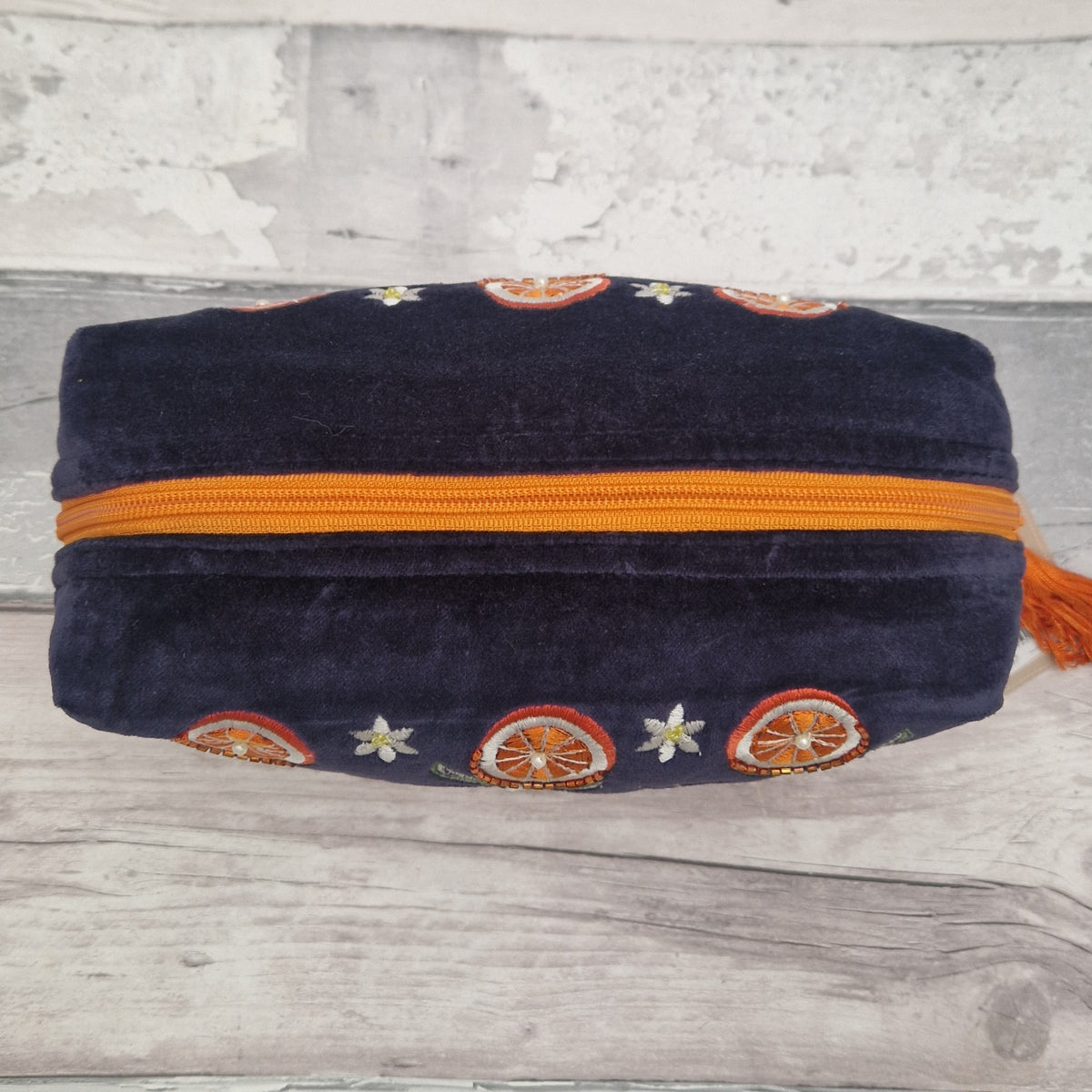 A navy blue velvet zippered pouch with orange and white embroidered citrus slice and flower patterns, resting on a light gray wooden surface.