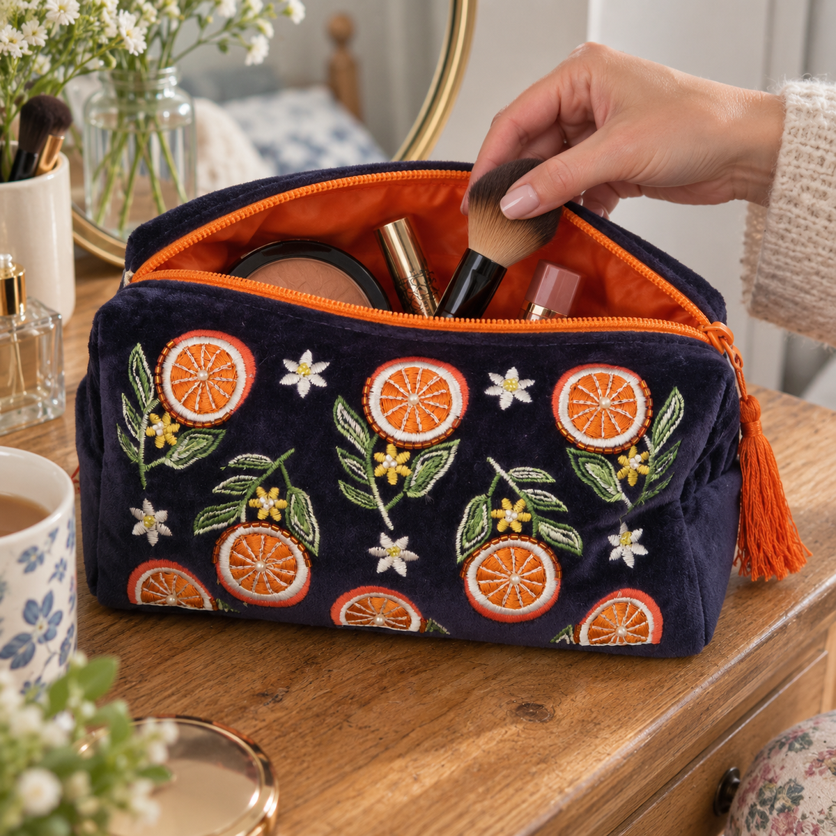 A navy blue velvet cosmetic pouch with embroidered orange slices, green leaves, and small white flowers, shown open on a wooden table with makeup brushes and cosmetics inside. A hand is visible reaching into the pouch.