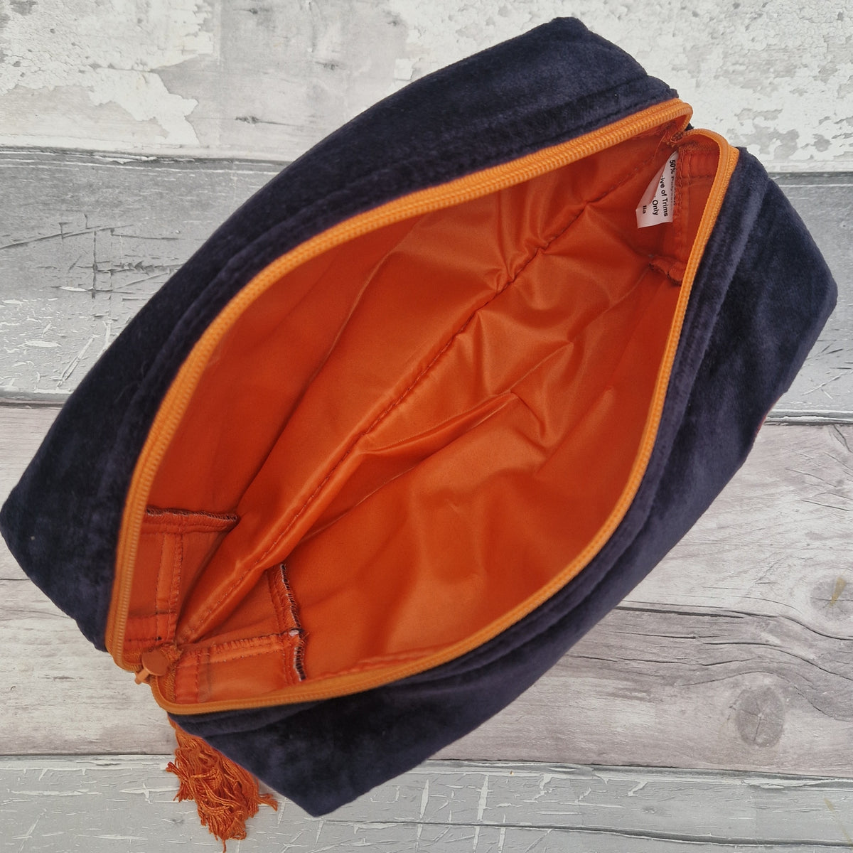 A navy blue velvet cosmetic pouch shown open, revealing a bright orange interior lining and zipper trim, placed on a light gray wood plank surface.