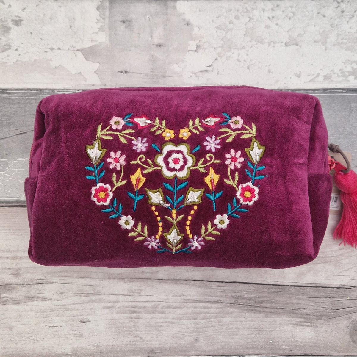 A deep magenta velvet cosmetic pouch featuring a colorful embroidered floral heart design on the front, resting on a rustic gray wooden surface.