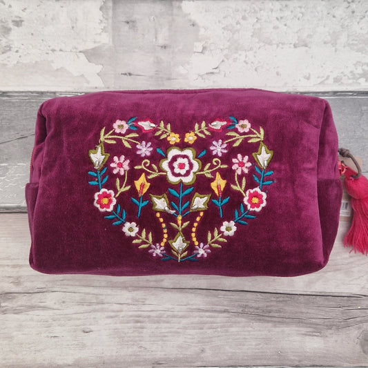 A deep magenta velvet cosmetic pouch featuring a colorful embroidered floral heart design on the front, resting on a rustic gray wooden surface.