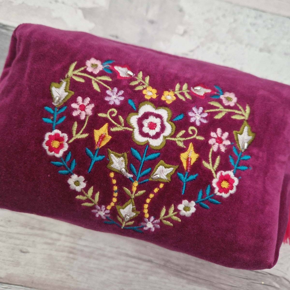 A velvet magenta washbag featuring a colorful embroidered floral heart design in the center, placed on a light gray wooden surface.