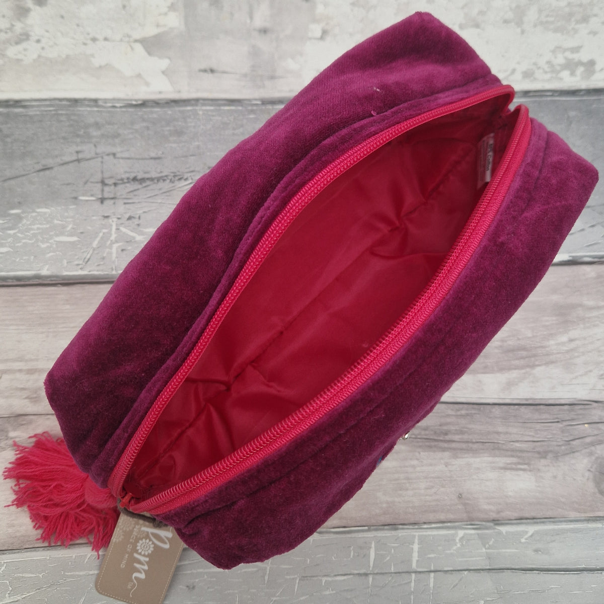 A deep magenta velvet cosmetic pouch with a matching pink zipper and tassel, shown open to reveal a pink interior. A small paper tag is attached near the tassel, and the pouch is photographed on a rustic gray wood background.