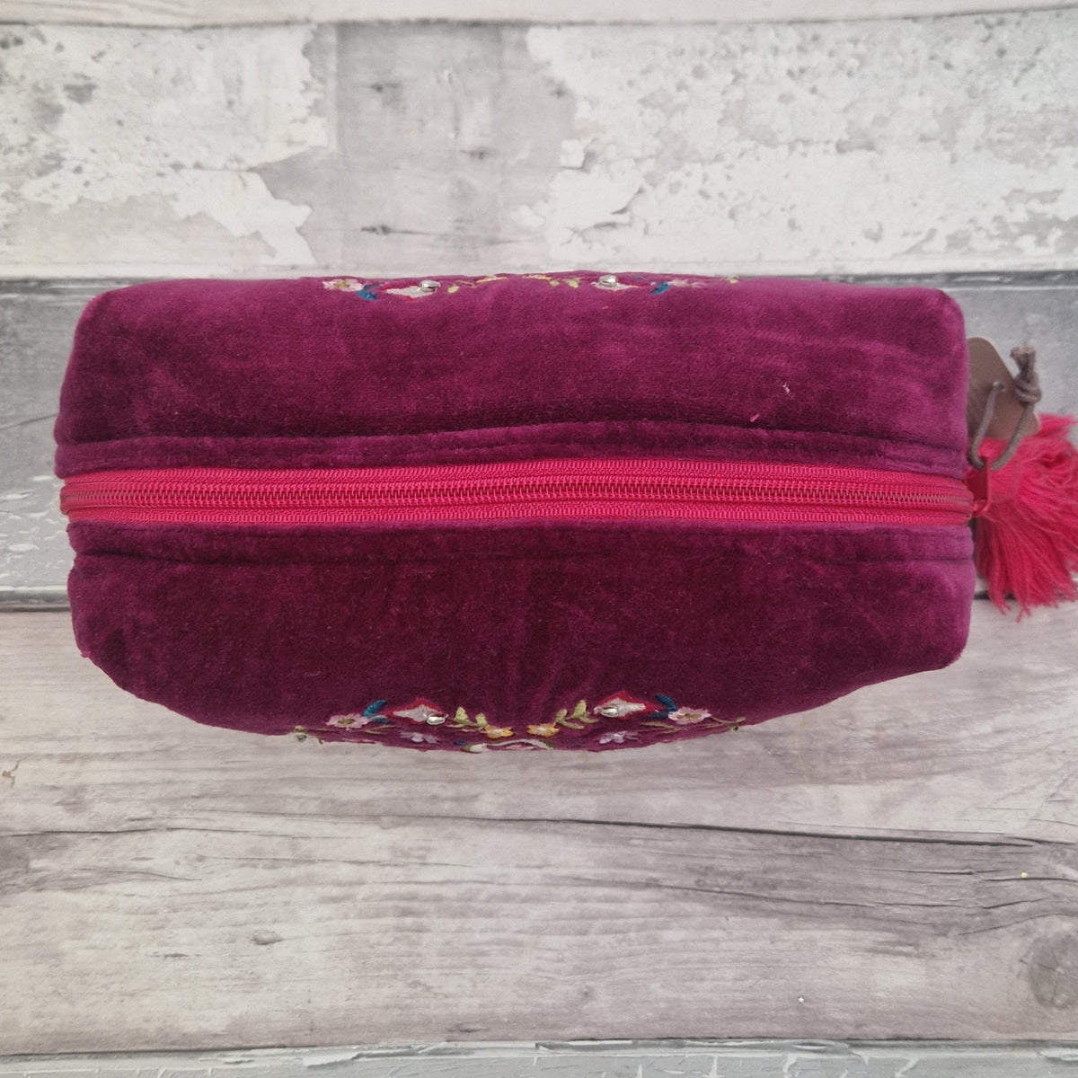A deep magenta velvet zippered pouch with colorful floral embroidery on the front and a matching tassel zipper pull, resting on a rustic gray wooden surface.