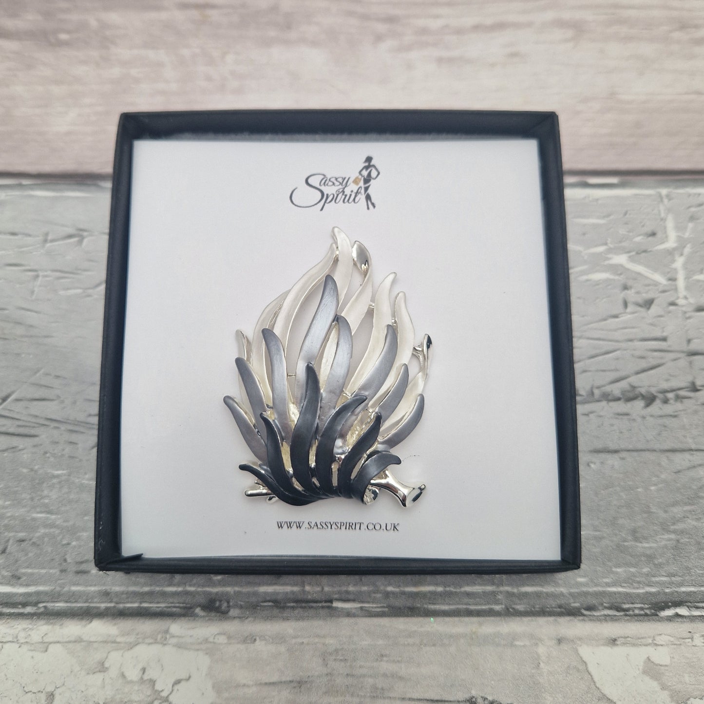 Silver and black fire flames brooch on a branded card with a wooden background