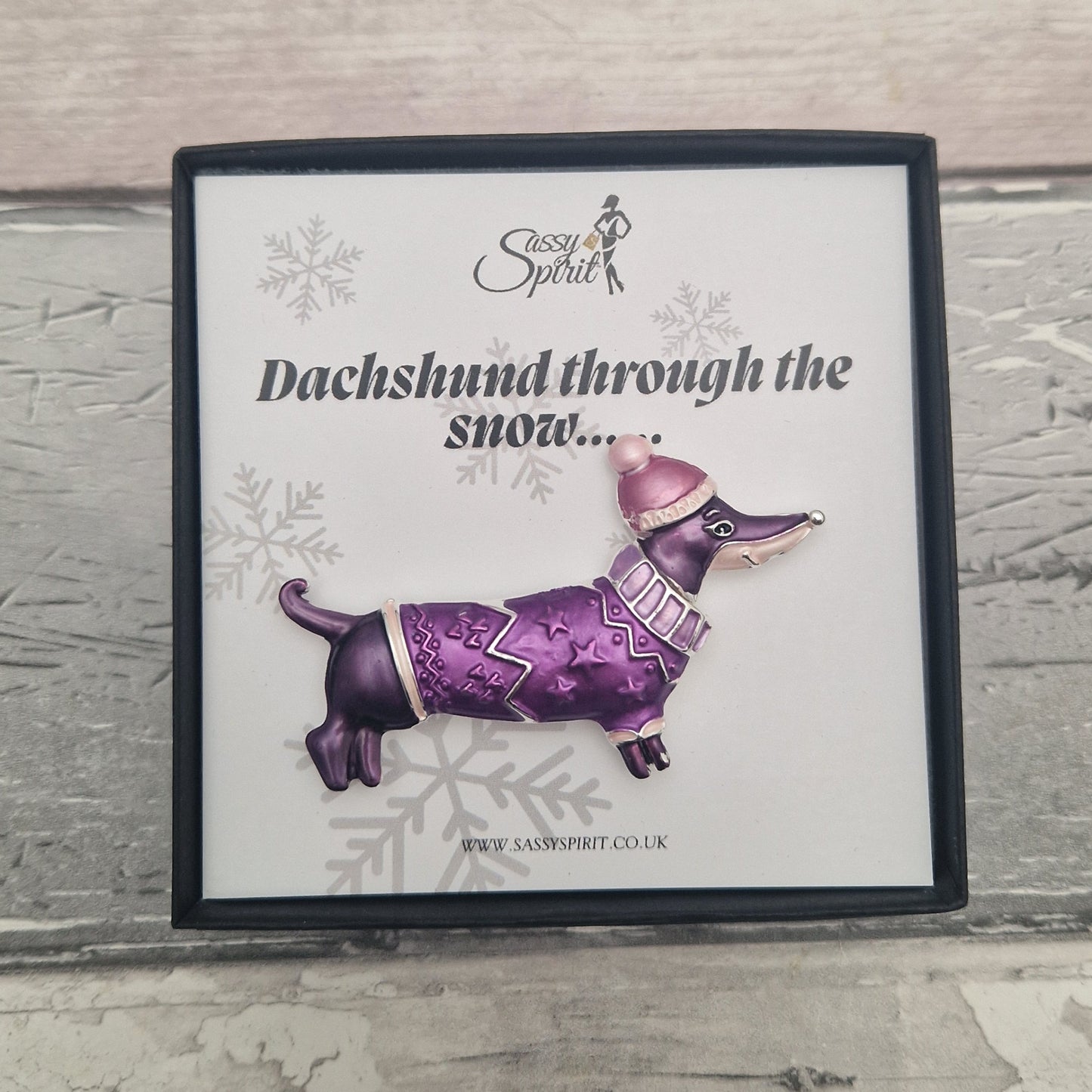 Purple dachshund brooch with a pink hat on a card with snowflake designs and text.