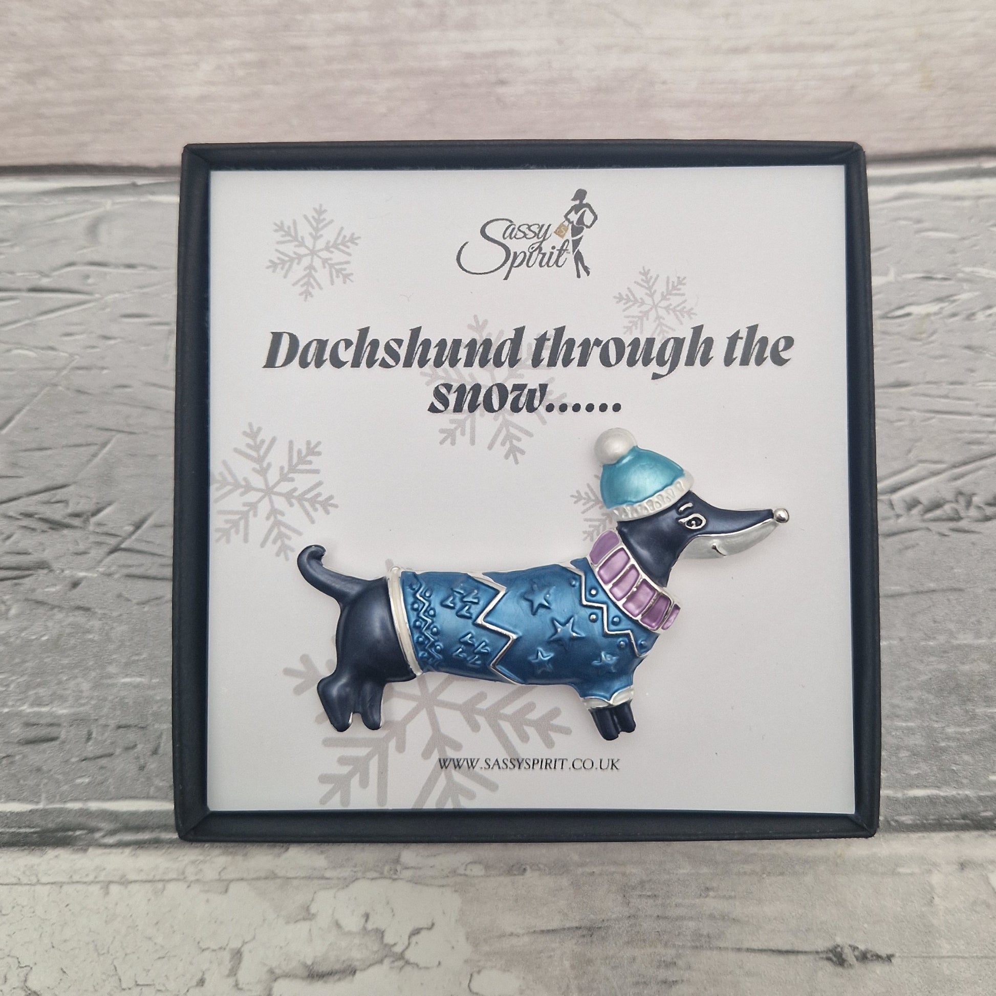 Pin with a dachshund wearing a blue sweater and hat on a decorative card with snowflake designs.