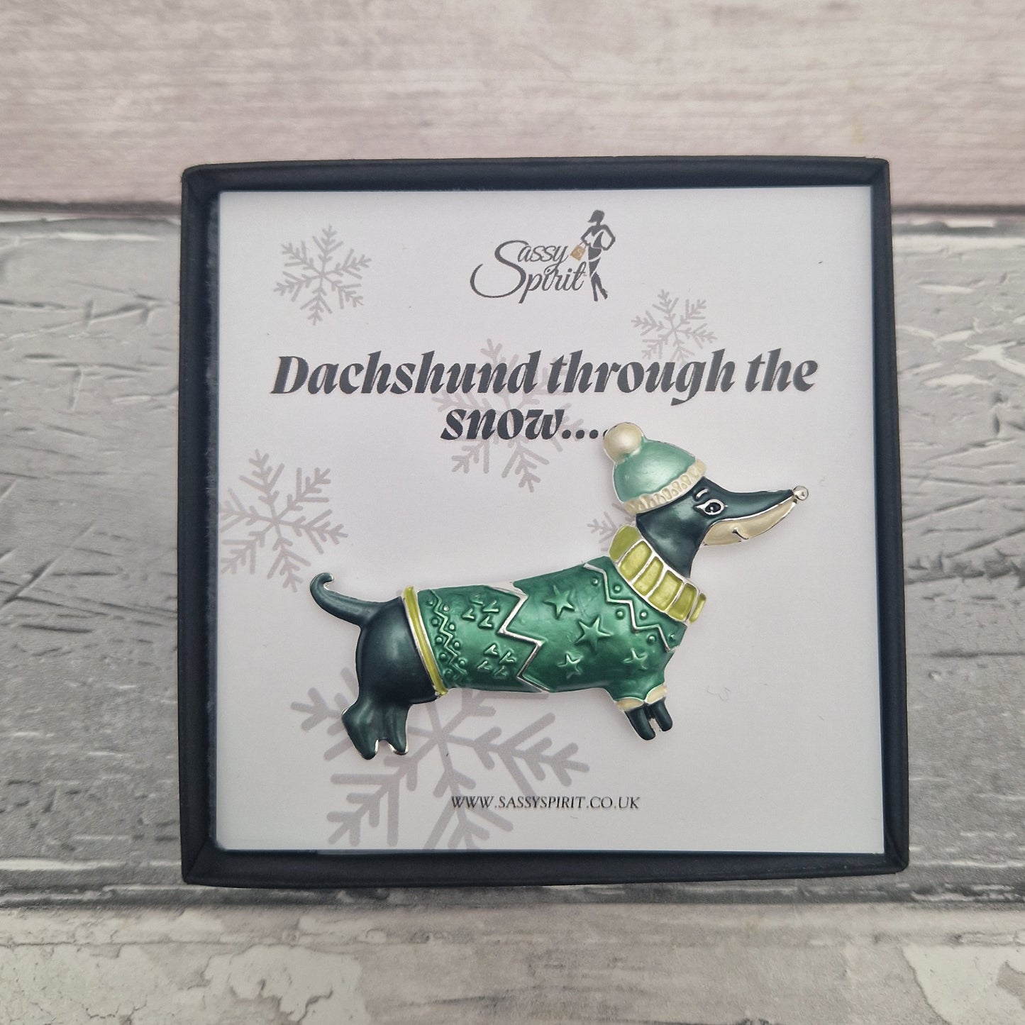 Brooch of a dachshund in a green sweater and hat on a branded card with snowflake designs.
