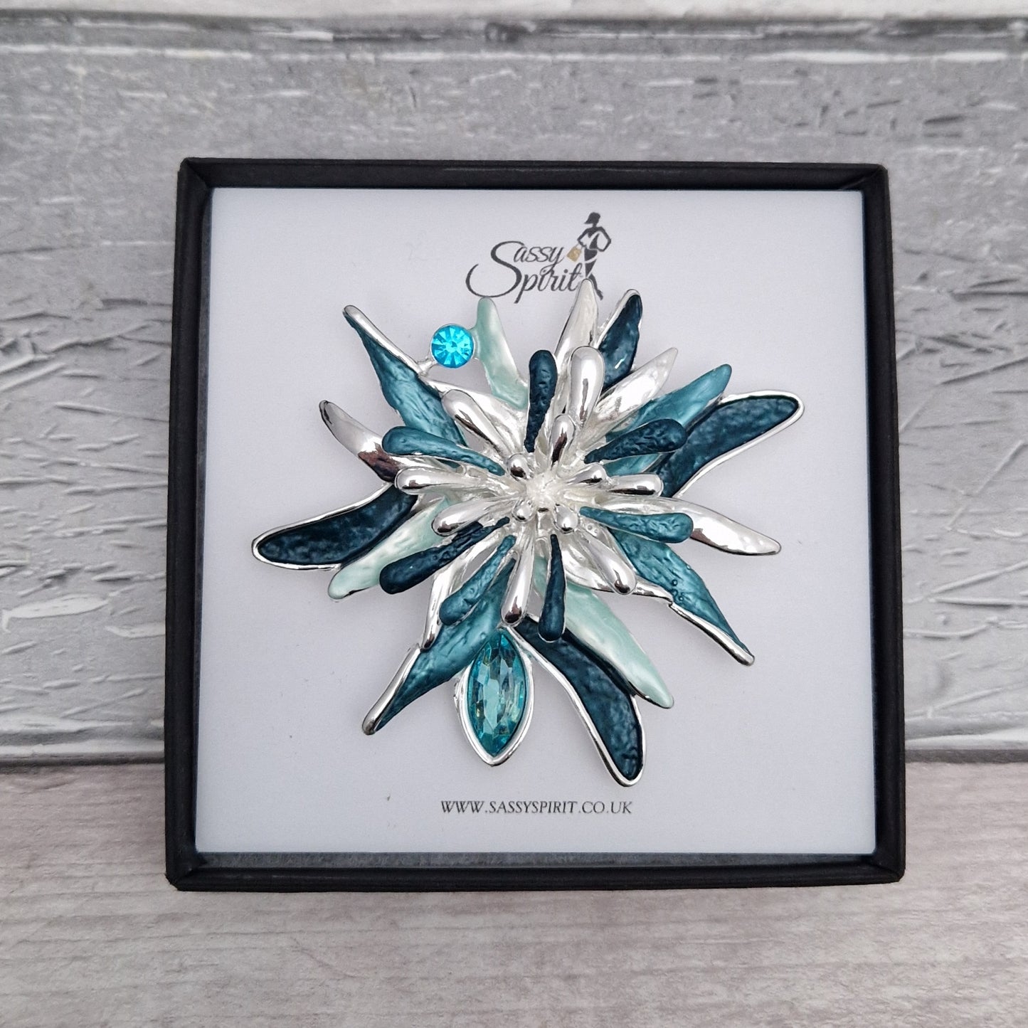 Silver and blue flower-shaped brooch on a branded display card with a gray background