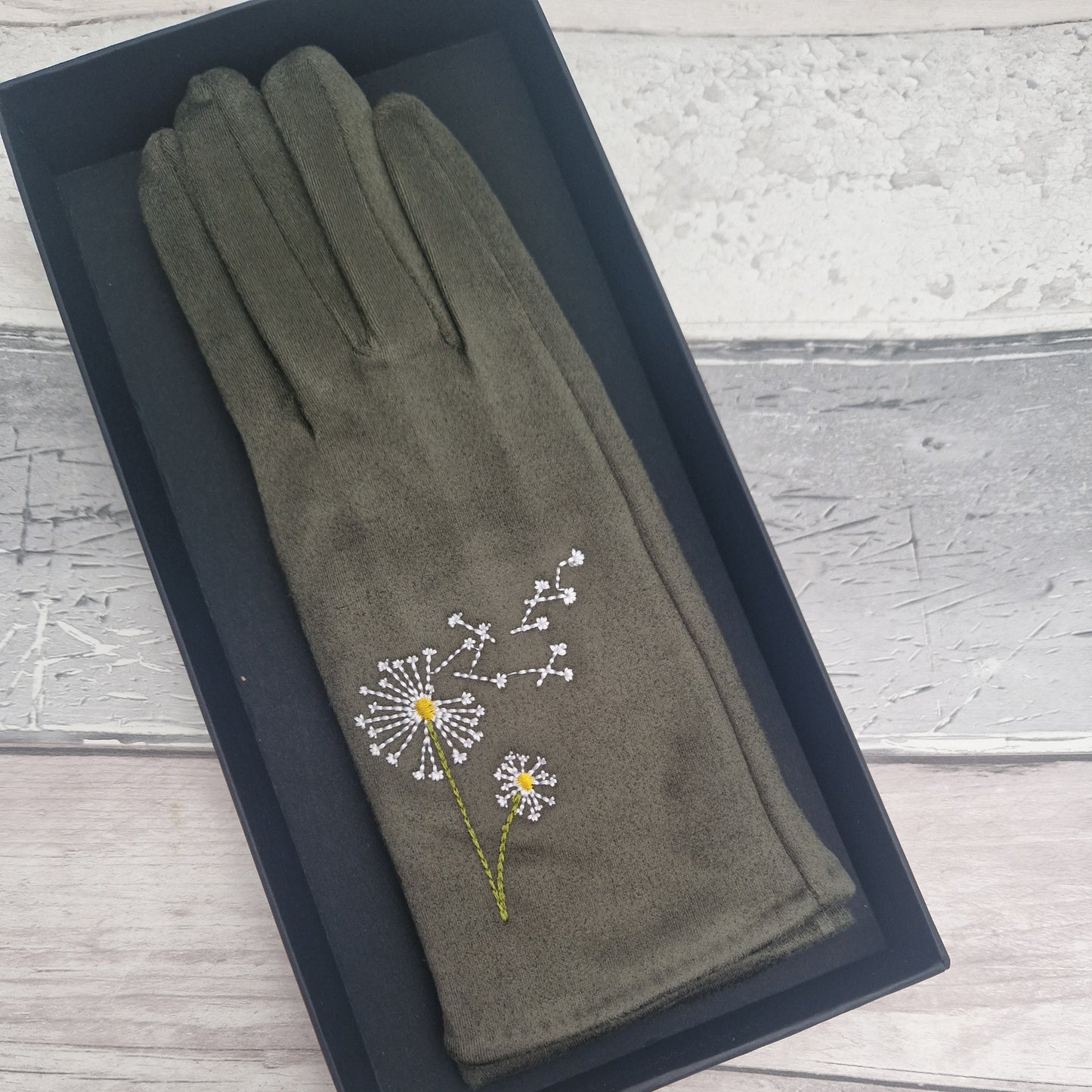 Green glove with dandelion embroidery in a black box on a wooden surface