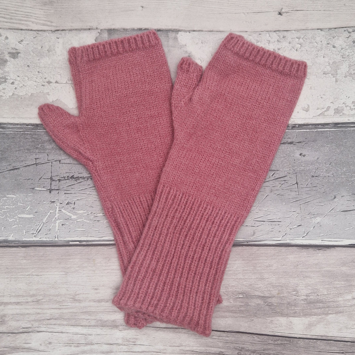 A pair of dusty rose pink knit fingerless gloves laid flat on a rustic, light gray weathered wood plank background.
