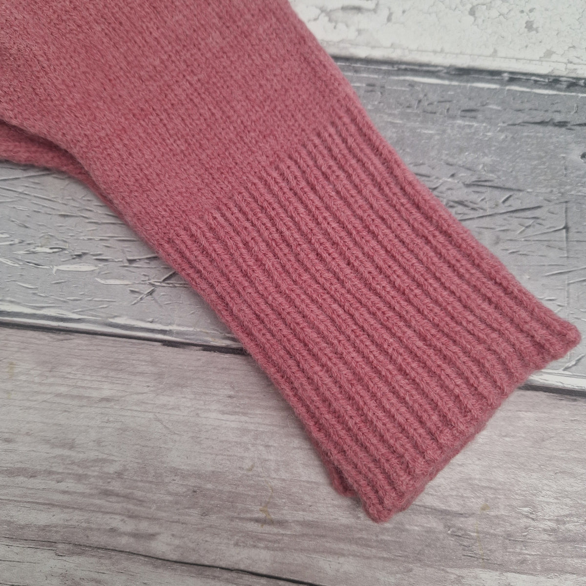 A close-up of a dusty rose pink ribbed knit sleeve or cuff laid diagonally on a rustic, weathered gray wooden surface.