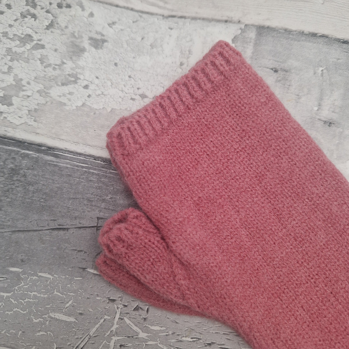 A single dusty rose pink knit glove or mitten with a ribbed cuff, laid on a distressed gray wooden surface.