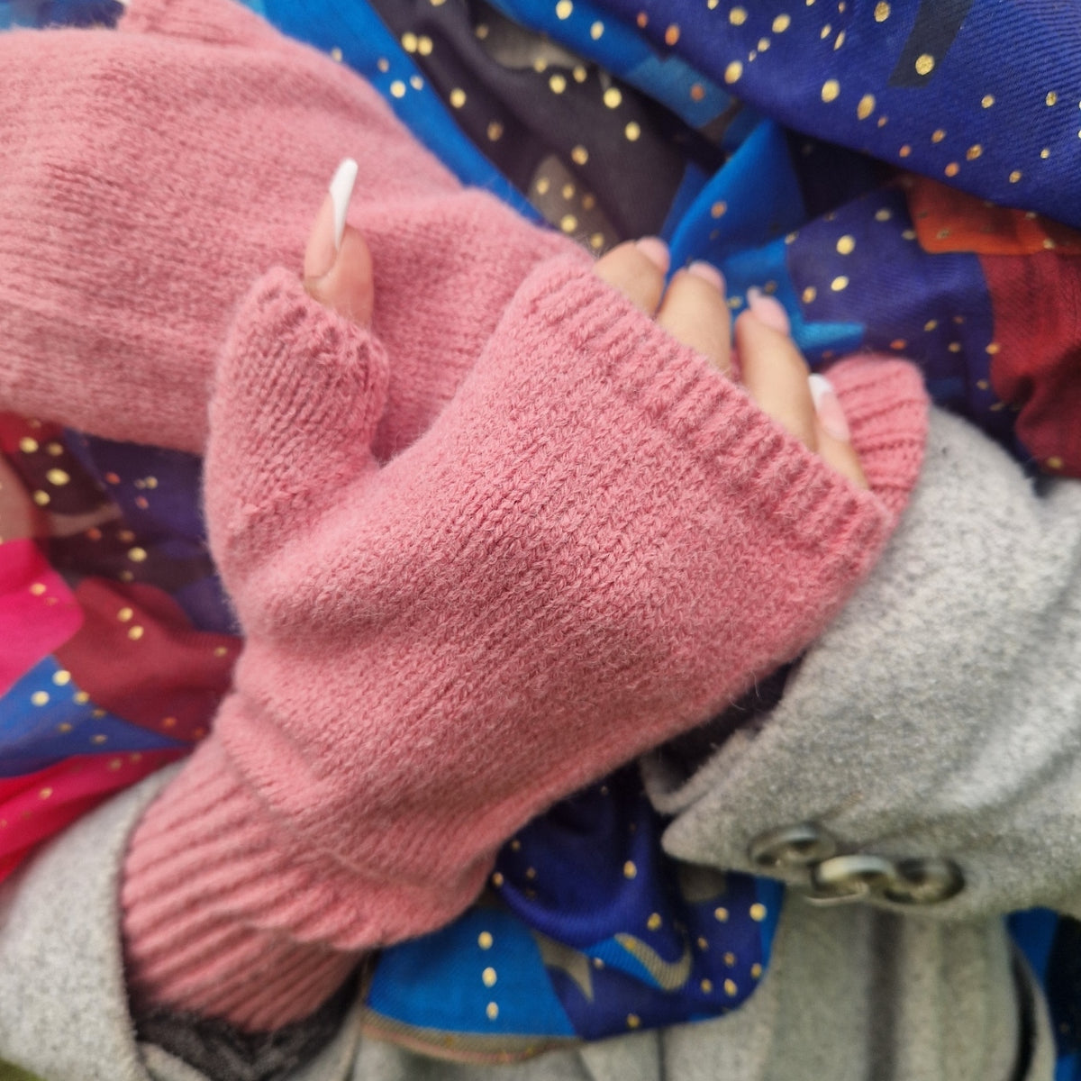 A close-up of a person wearing a pair of pink fingerless knit gloves, holding a gray coat and colorful patterned fabric.